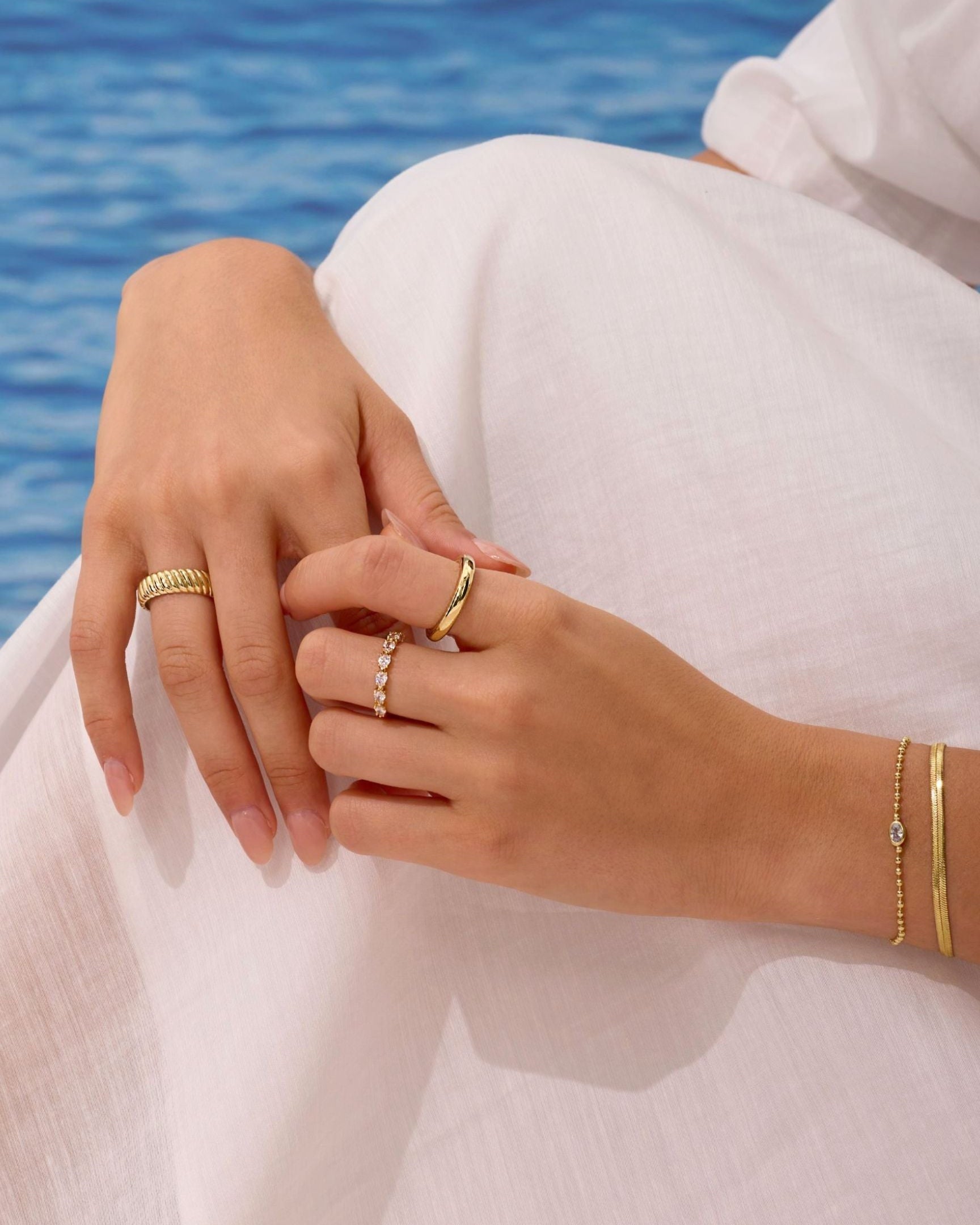 Close-up of hands with gold rings and bracelets against a blurred blue water background