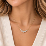 A close-up of a woman wearing a delicate gold necklace with a curved row of clear gemstones at the center, paired with a cream-colored knit top.