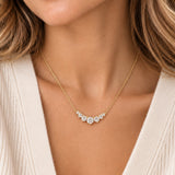 A close-up of a woman wearing a delicate gold necklace with a curved row of clear gemstones at the center, paired with a cream-colored knit top.