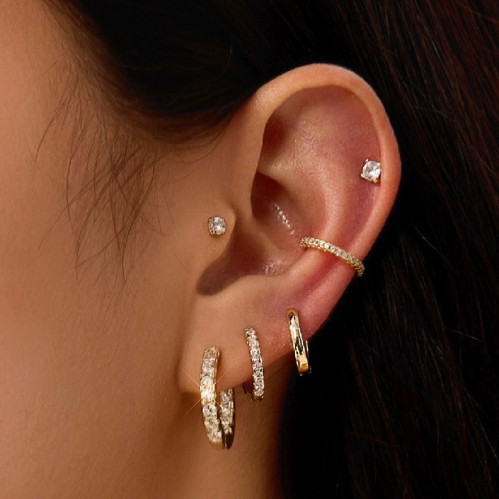A close-up of a person's ear showcasing multiple gold and silver ear piercings, including hoop earrings, a cartilage stud, a helix stud, a lobe stud, and a curved barbell cartilage piercing.