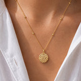 A close-up of a gold necklace with a textured circular pendant and small bead accents on the chain, worn on a model's neck over a white collared shirt.