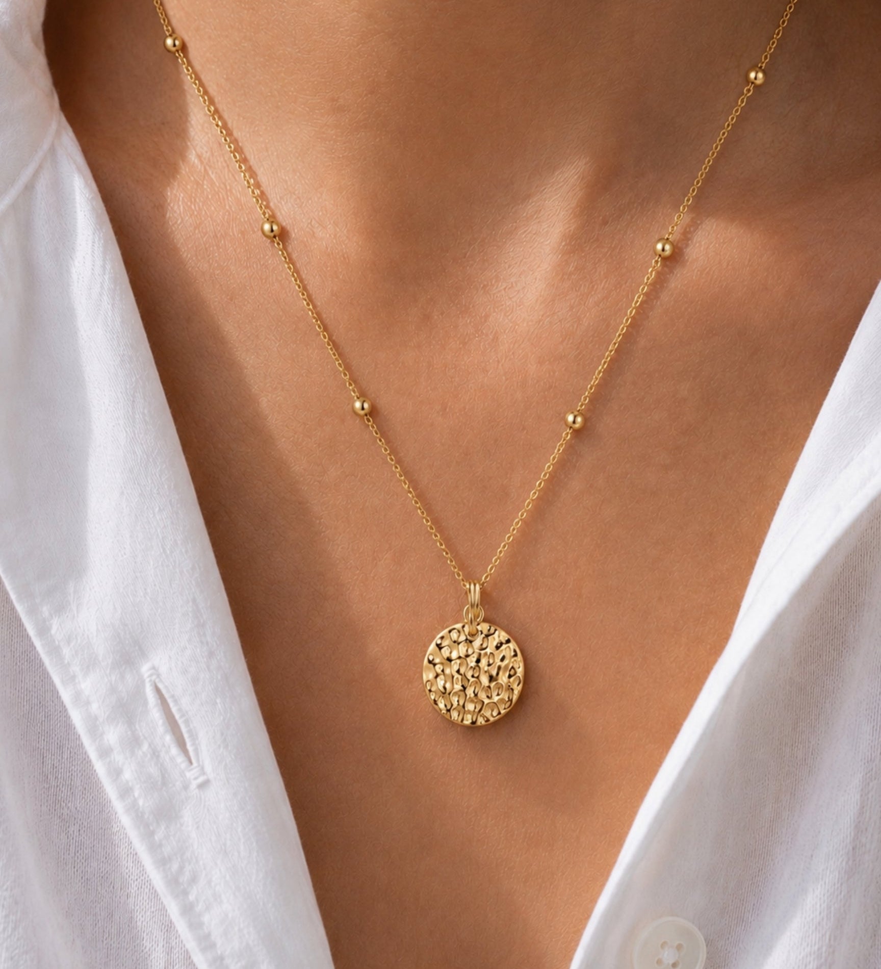 A close-up of a gold necklace with a textured circular pendant and small bead accents on the chain, worn on a model's neck over a white collared shirt.