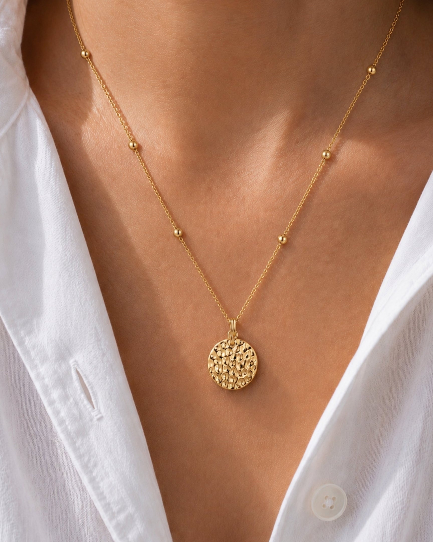 A close-up of a gold necklace with a textured circular pendant and small bead accents on the chain, worn on a model's neck over a white collared shirt.