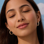 Close-up of a woman with closed eyes wearing gold hoop earrings against a blue sky.