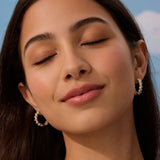 Close-up of a woman with closed eyes wearing gold hoop earrings against a blue sky.