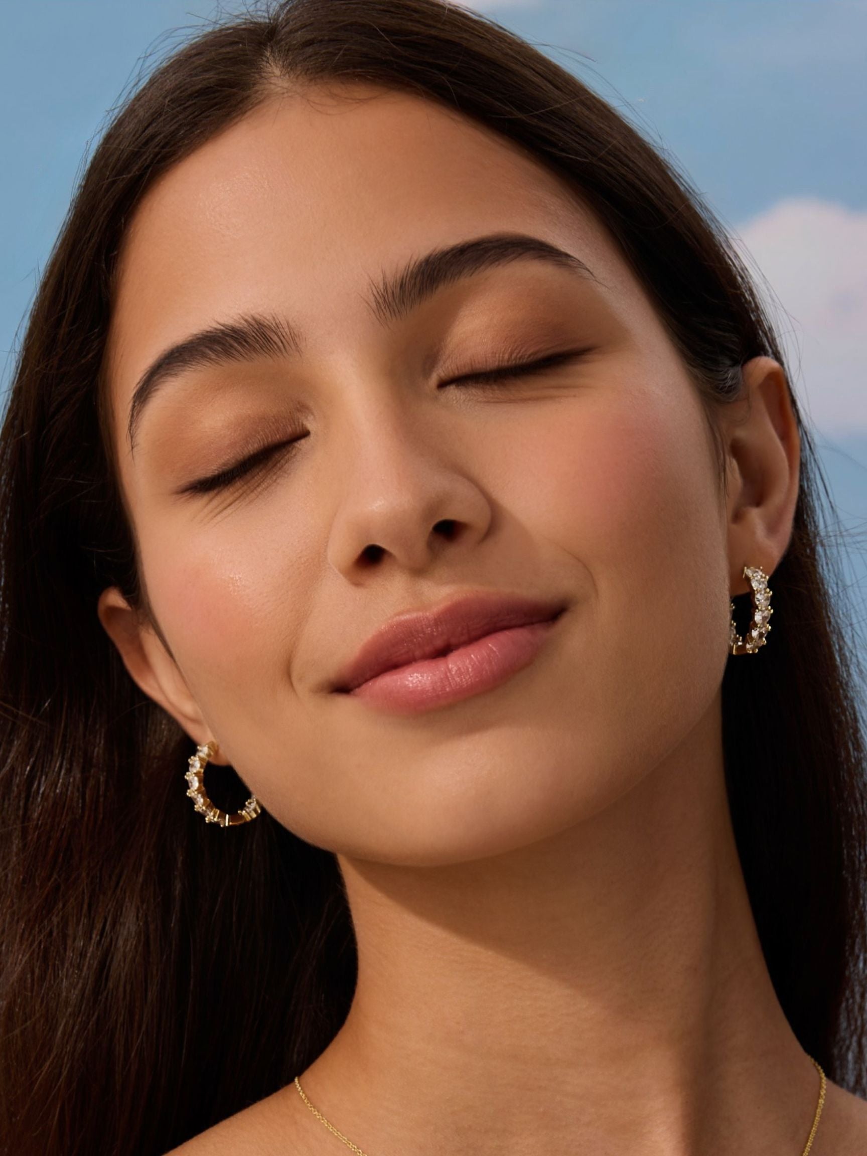 Close-up of a woman with closed eyes wearing gold hoop earrings against a blue sky.